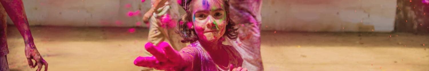 A child with colorful powder on their face and hands, smiling and reaching forward during a vibrant outdoor celebration.