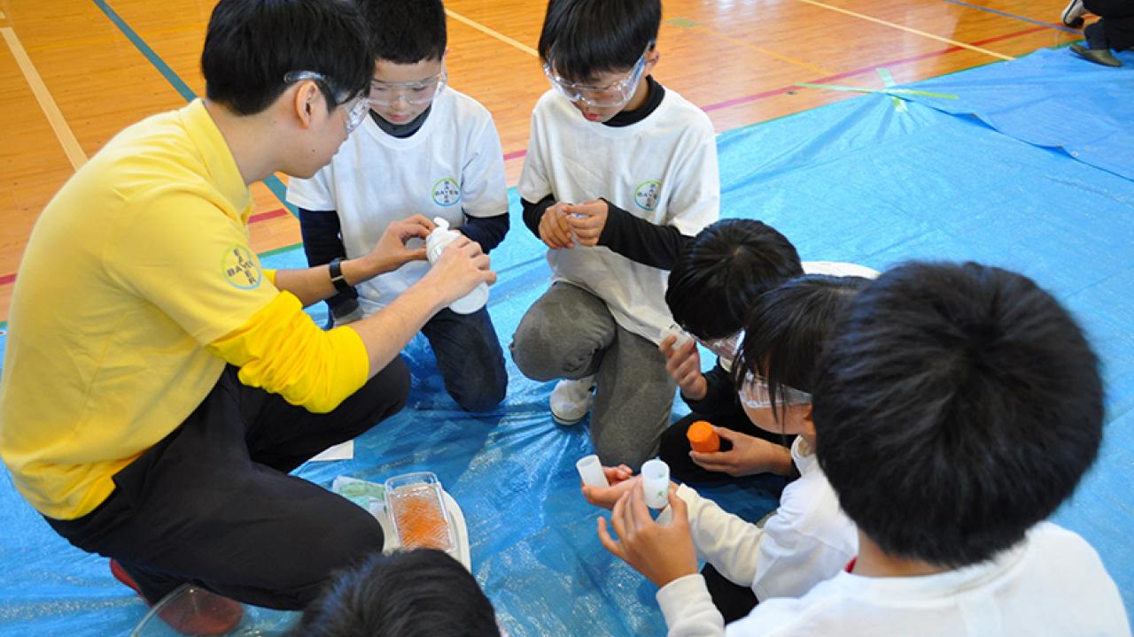 安全ゴーグルを着けた子供たちのグループが大人の周りに集まり、屋内の青い防水シートの上でボトルを使った科学実験に取り組んでいます。