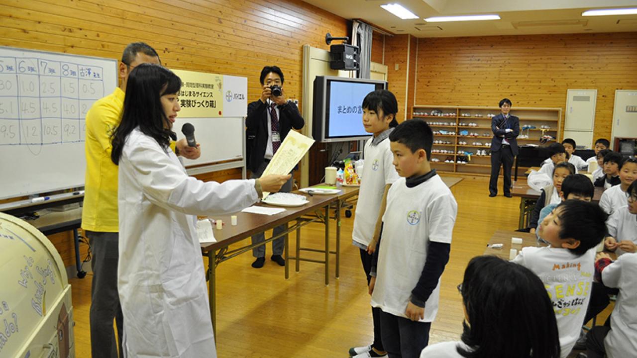 白衣を着た人物が教室で子供たちに話しかけています。左側には数字が書かれたボードがあり、数人がそのやり取りを見守っています。