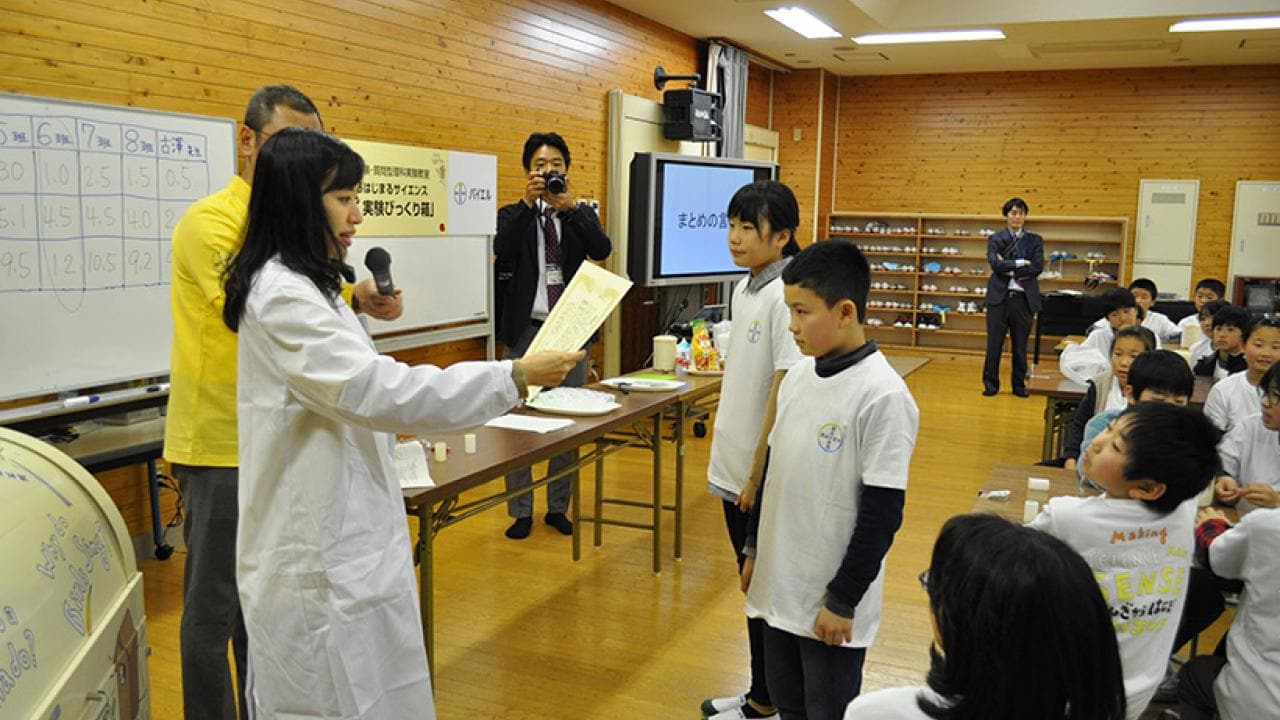 白衣を着た人物が教室で子供たちに話しかけています。左側には数字が書かれたボードがあり、数人がそのやり取りを見守っています。