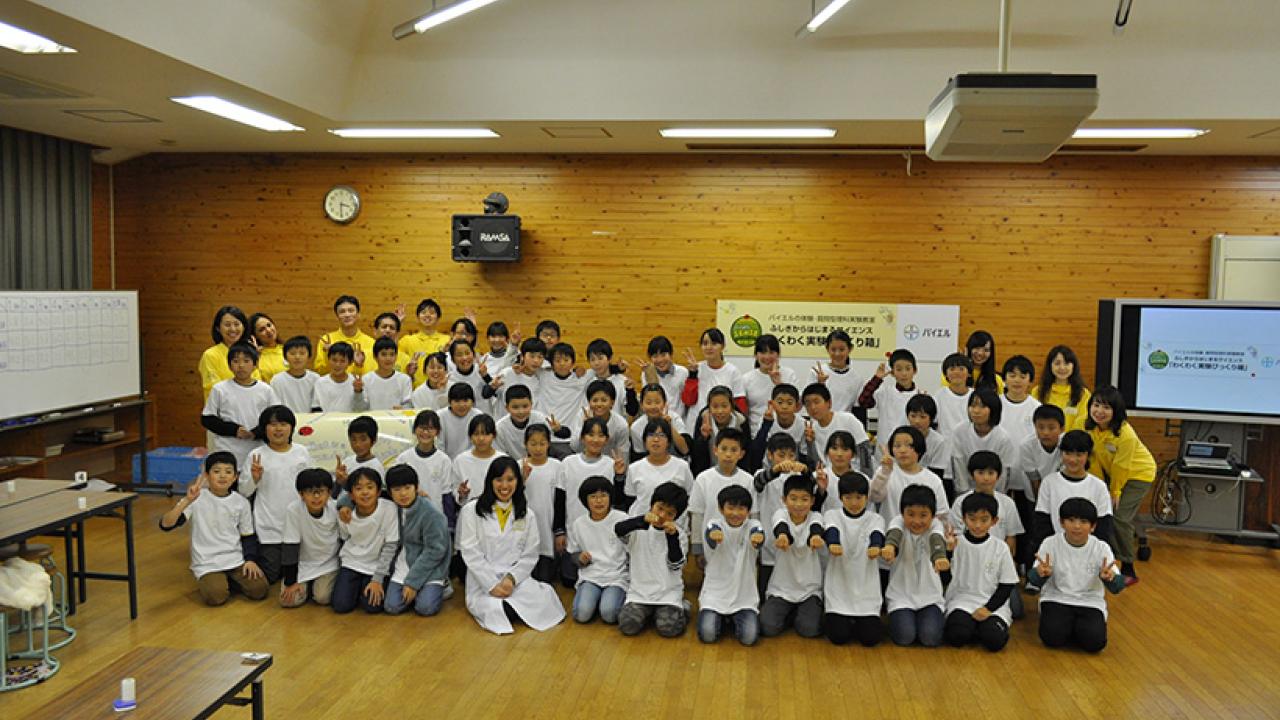 白いシャツと黄色いベストを着た大勢の子供と大人が、木製の壁とプレゼンテーションスクリーンのある部屋で写真を撮っています。