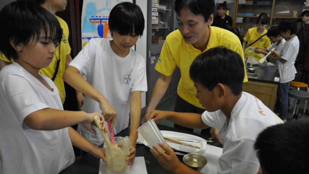 教室では、子どもたちと先生がカジュアルな制服を着て、容器の中で物質を混ぜる理科の実験を行っています。