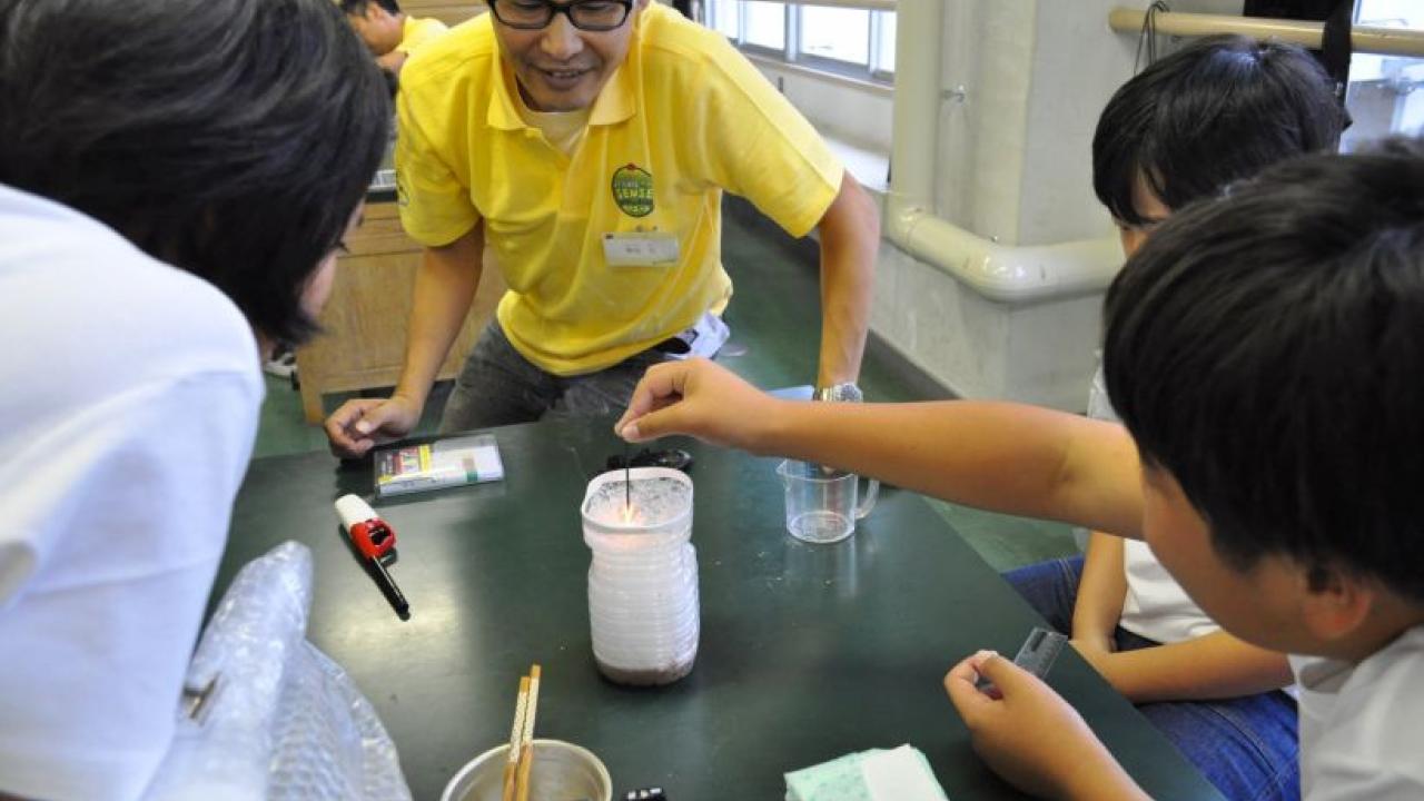 子どもと大人が、ペットボトルやビーカーなどさまざまな材料を使ってテーブルで科学実験を行っています。