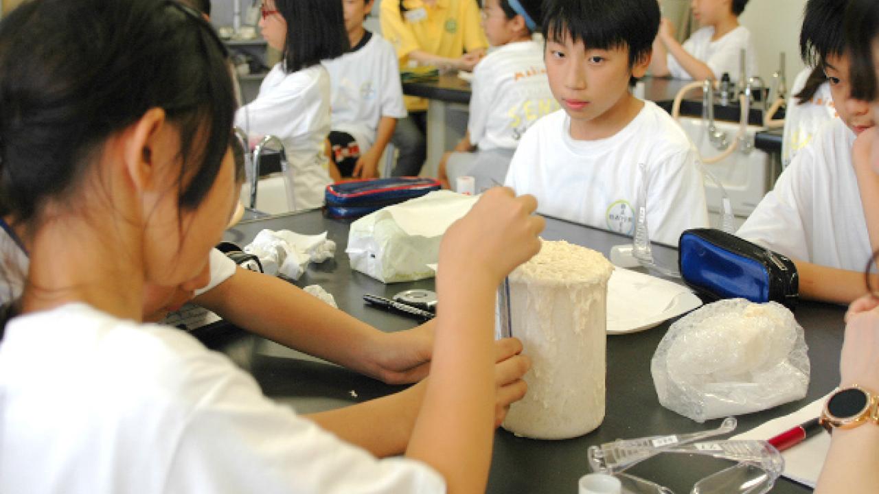 教室の子どもたちが、テーブルの上に置かれたさまざまな材料を使って科学プロジェクトに取り組んでいます。