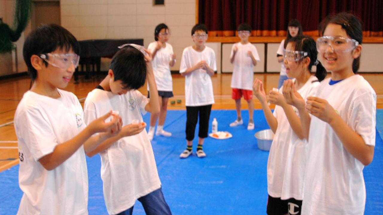 保護ゴーグルと白いTシャツを着た子どもたちが体育館の青い防水シートの上に立ち、実践的な活動に取り組んでいます。