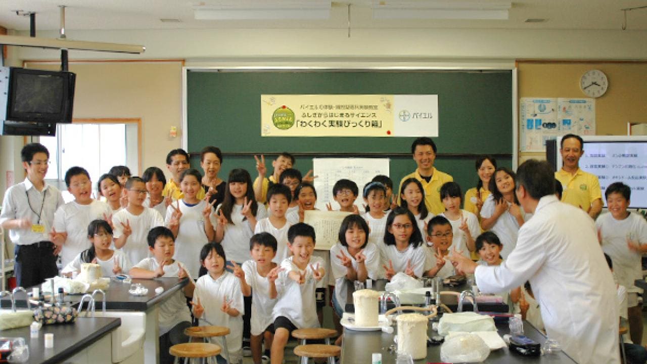 教室にいる子供と大人のグループが写真のポーズをとっています。白衣を着た人が前に立っています。
