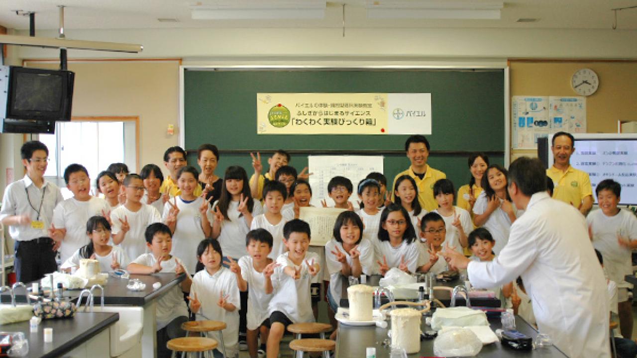 教室にいる子供と大人のグループが写真のポーズをとっています。白衣を着た人が前に立っています。