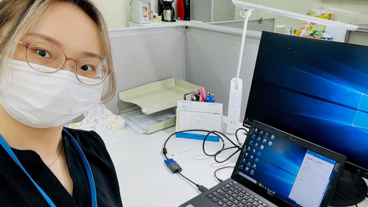 マスクとメガネを着用した人物が、オフィスの環境で、机、ノートパソコン、モニターを背景に自撮り写真を撮影しています。