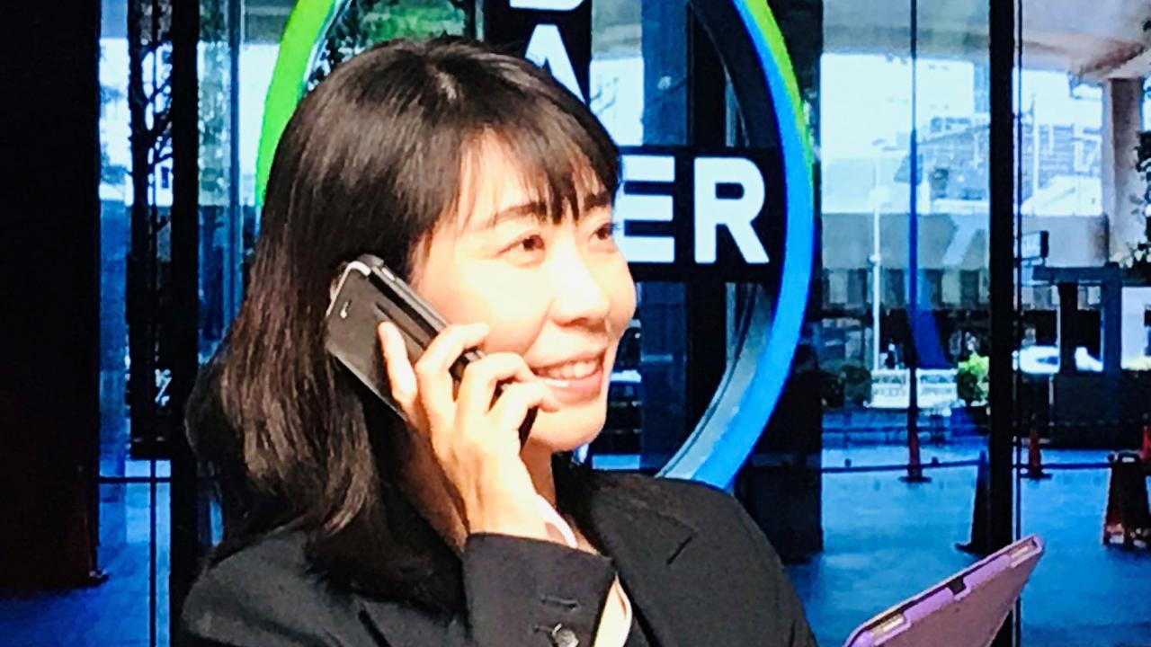 ビジネススーツを着た女性がタブレットを持ちながらスマートフォンで通話しており、背景にはガラス張りの建物の入り口と円形の看板がある。