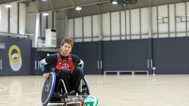 屋内スポーツコートで車椅子に乗っている人と、その近くにバレーボールがある。
