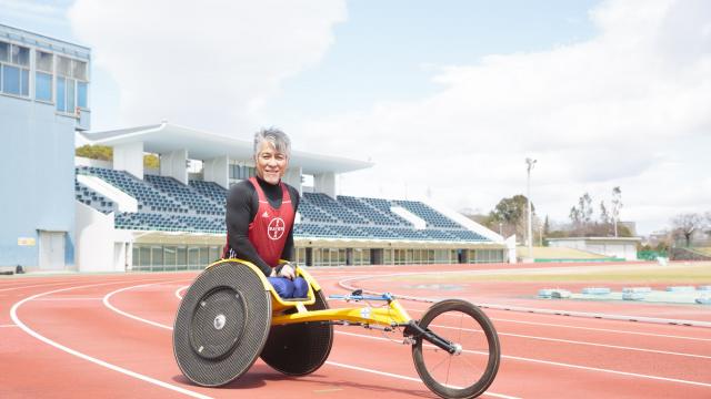 赤いジャージを着て、競技用車椅子に乗ってトラックを走る選手。曇り空の下、背景にスタジアムの座席が見える。