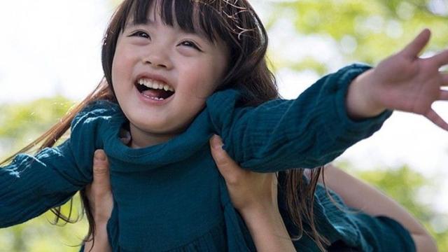 青いドレスを着た子供が両手を広げて抱き上げられ、屋外で満面の笑みを浮かべている。