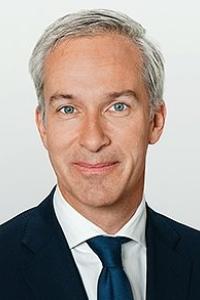 A man with grey hair, wearing a suit and blue tie, smiling against a neutral background.