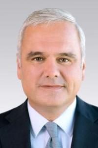 A man with short grey hair wearing a suit and tie, on a plain background.
