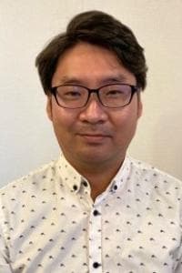 眼鏡をかけ、柄物のシャツを着た人が無地の背景に立っています。