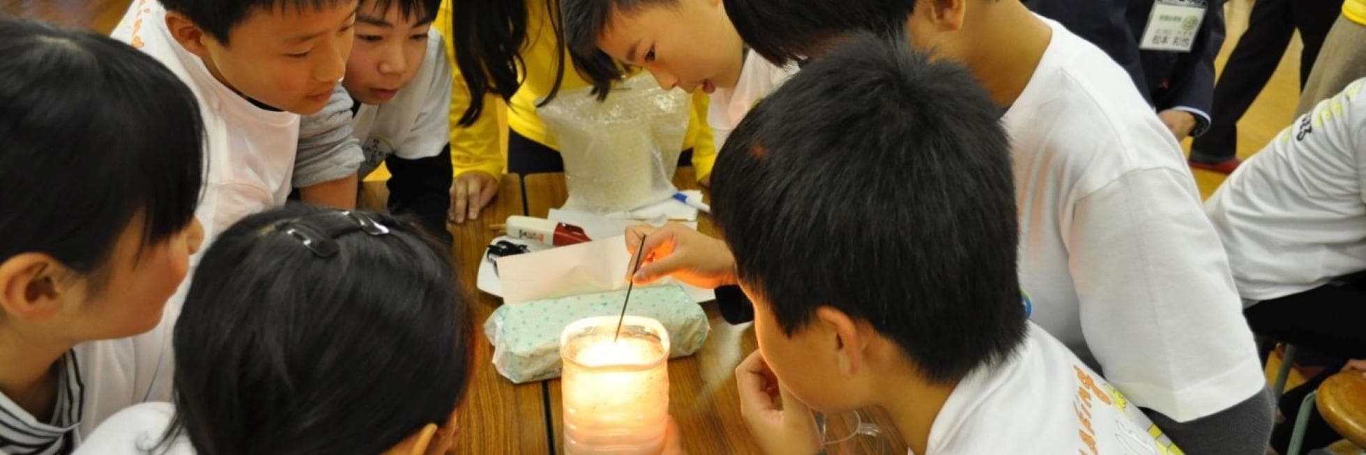 子どもたちはテーブルの周りに集まり、ビーカーの中に火のついたろうそくを入れる科学実験を観察したり、実験に参加したりしていました。